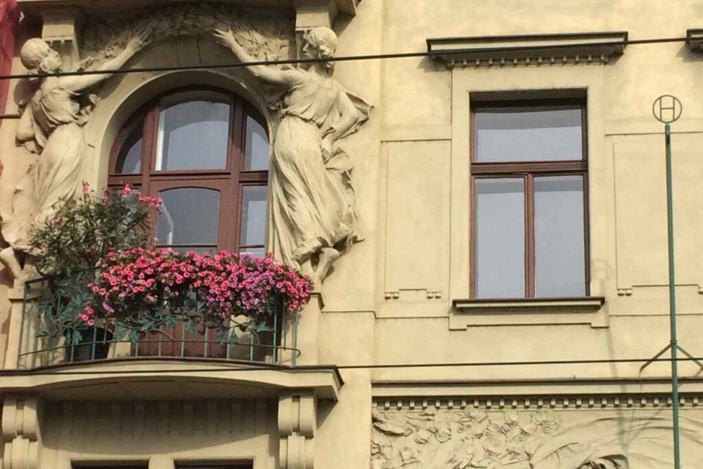 Two women sculpted on a facade in Prague, with some pink flowers on the balcony.